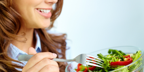 nasmejana brineta jede vitaminsku salatu viljuskom iz staklene cinije sa plavom kosuljom paradajz zelena salata paprika